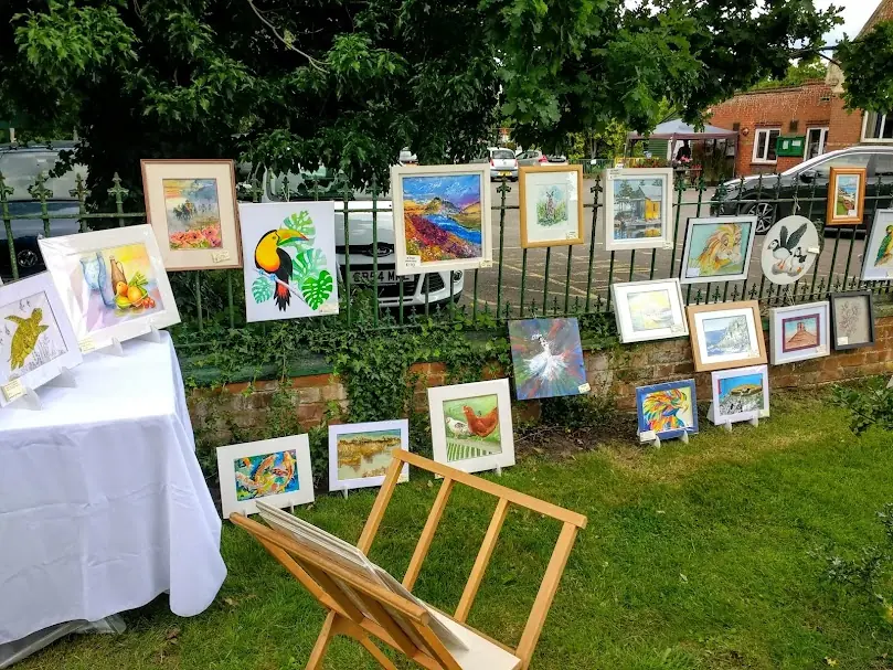 Framed paintings hung along garden railings in sunshine at the Pirbright exhibition